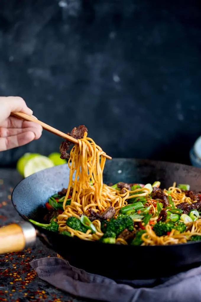 Side-on image of crispy chilli beef noodles with broccoli and peppers in a wok. 
The noodles are being lifted by some chopsticks held in a hand.
The wok is on a grey background with ingredients scattered around and a grey napkin in front of the wok.