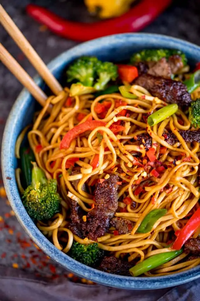 Overhead image of crispy chilli beef noodles with red pepper and broccoli in a blue bowl. There is a pair of chopsticks sticking out of the bowl.