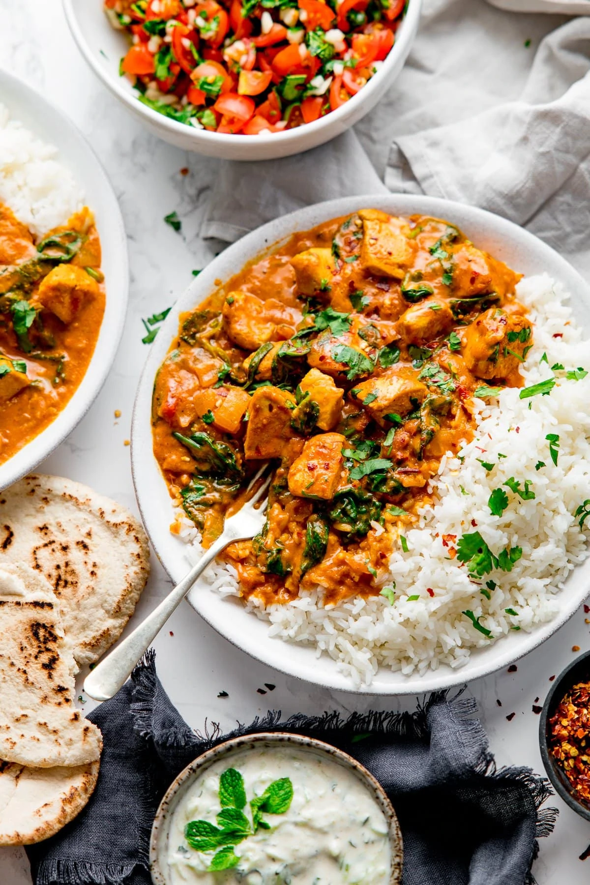 White bowl with chicken curry and rice with a fork digging into the curry. Ingredients and side dishes scattered around.