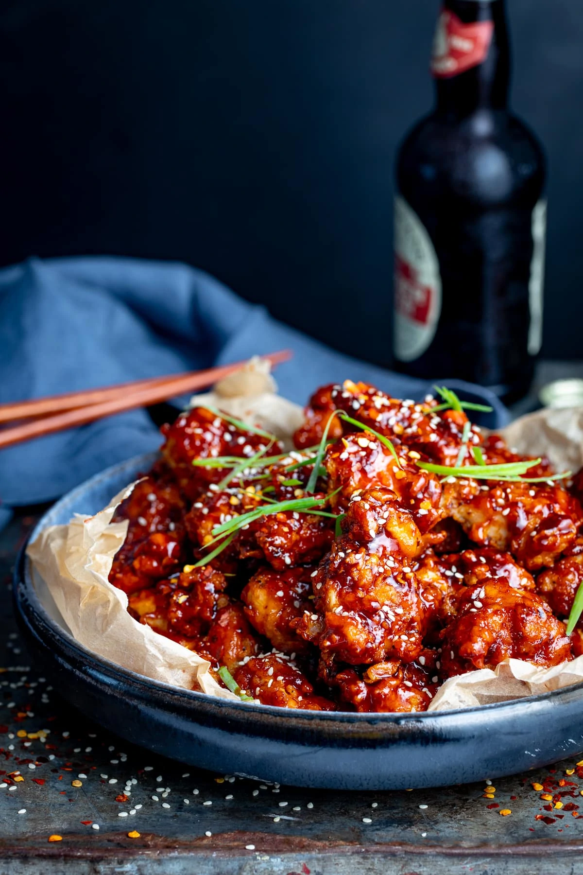 Korean fried chicken in a blue bowl against a dark background. Dark bottle and chopsticks in the background.