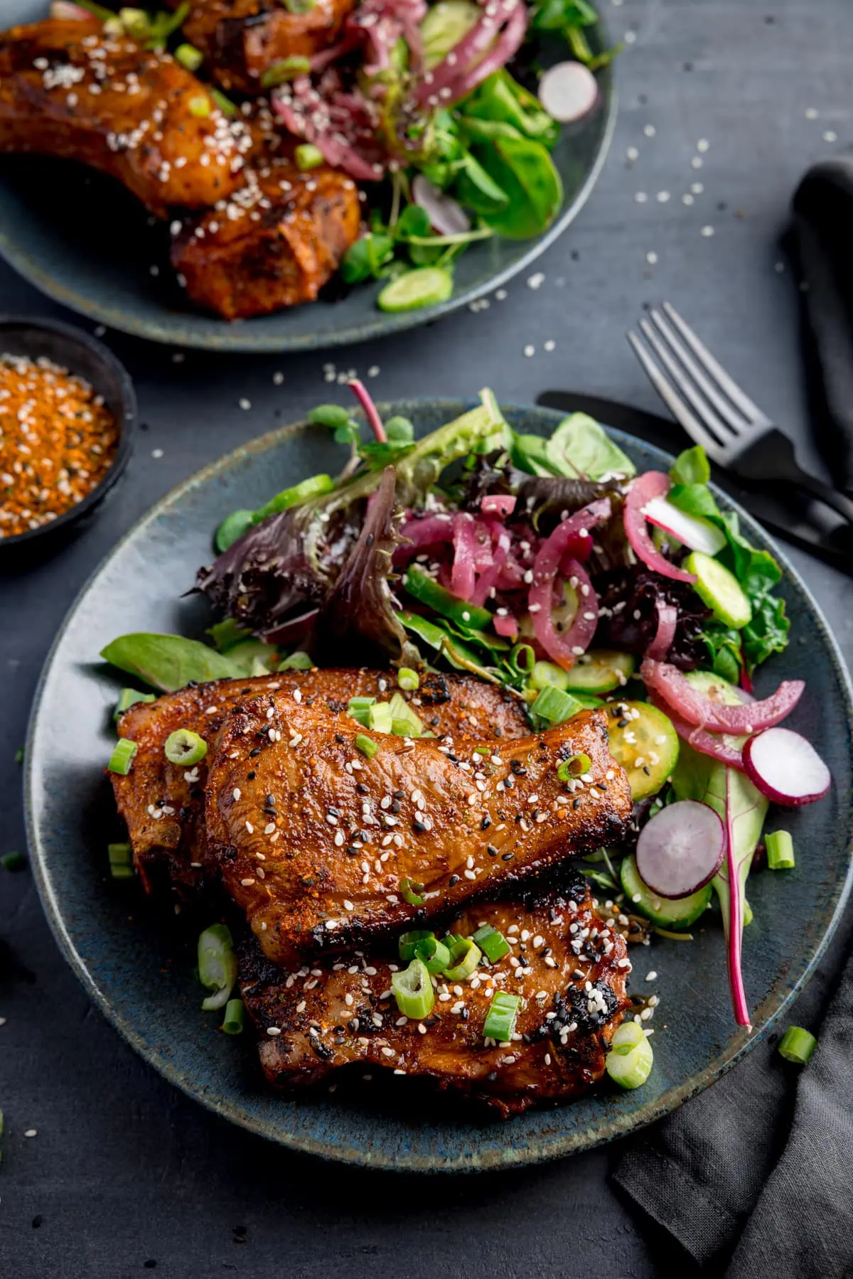 3 marinated and cooked lamb cutlets on a dark blue plate next to a side salad. The lamb cutlets are sprinkled with sesame seeds and chopped spring onions. The plate is on a dark grey background, next to a dark knife and fork and a further plate of lamb cutlets.