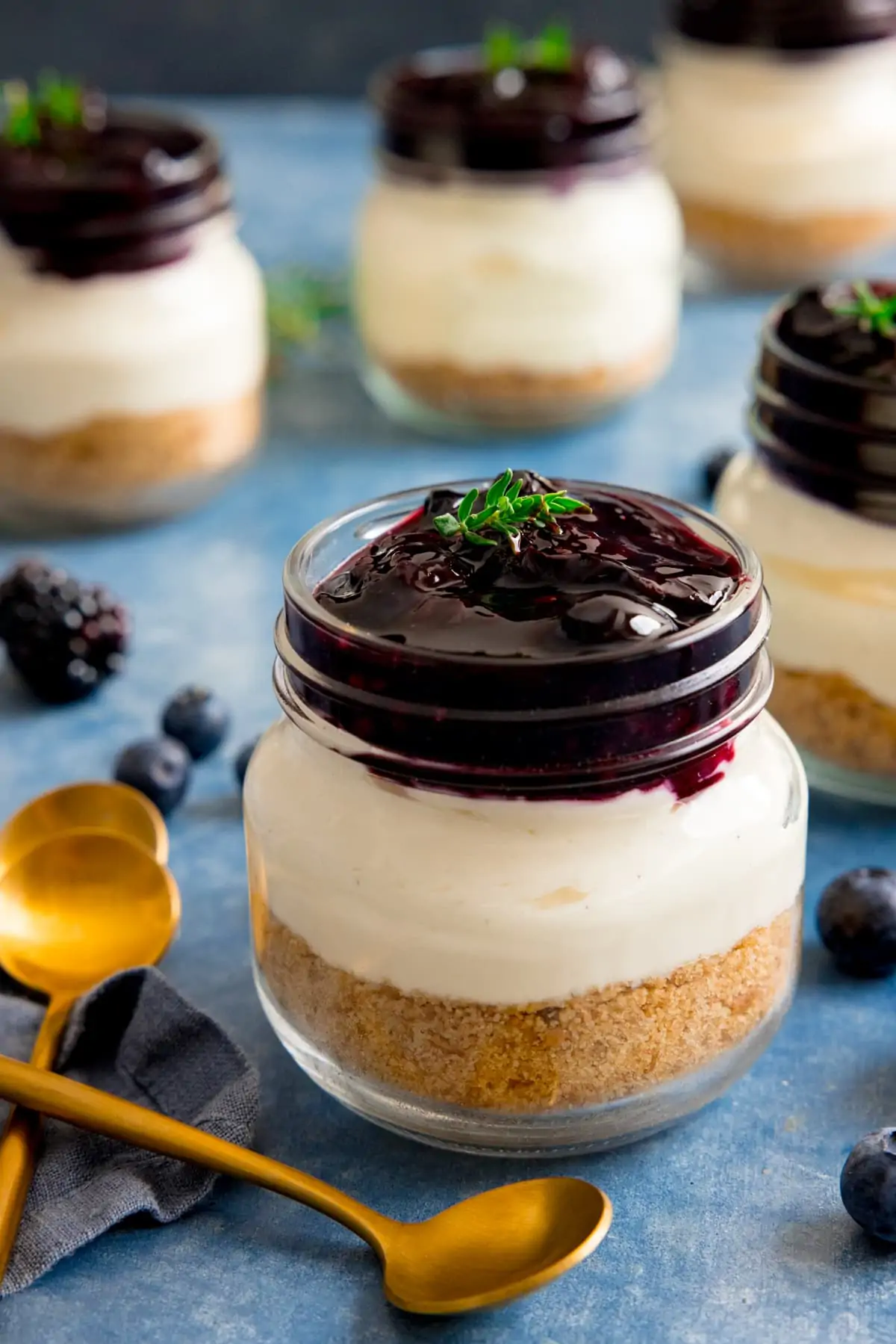 Mason jar berry cheesecake on a blue surface next to two gold teaspoons. Further mason jar cheesecakes in the background.