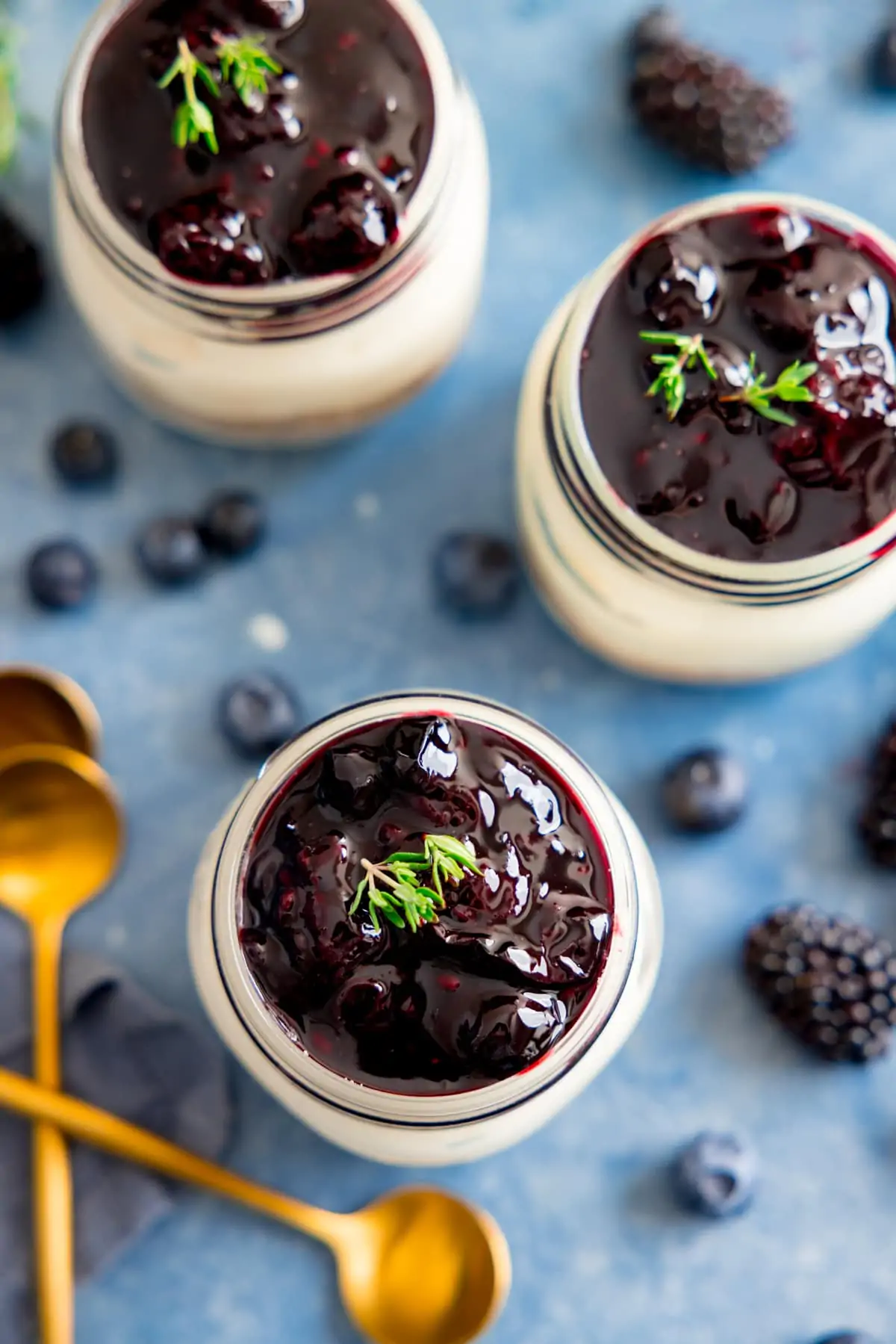 Overhead image of 3 mason jar individual berry cheesecakes on a blue background. Berries and gold teaspoons are next to the cheesecakes