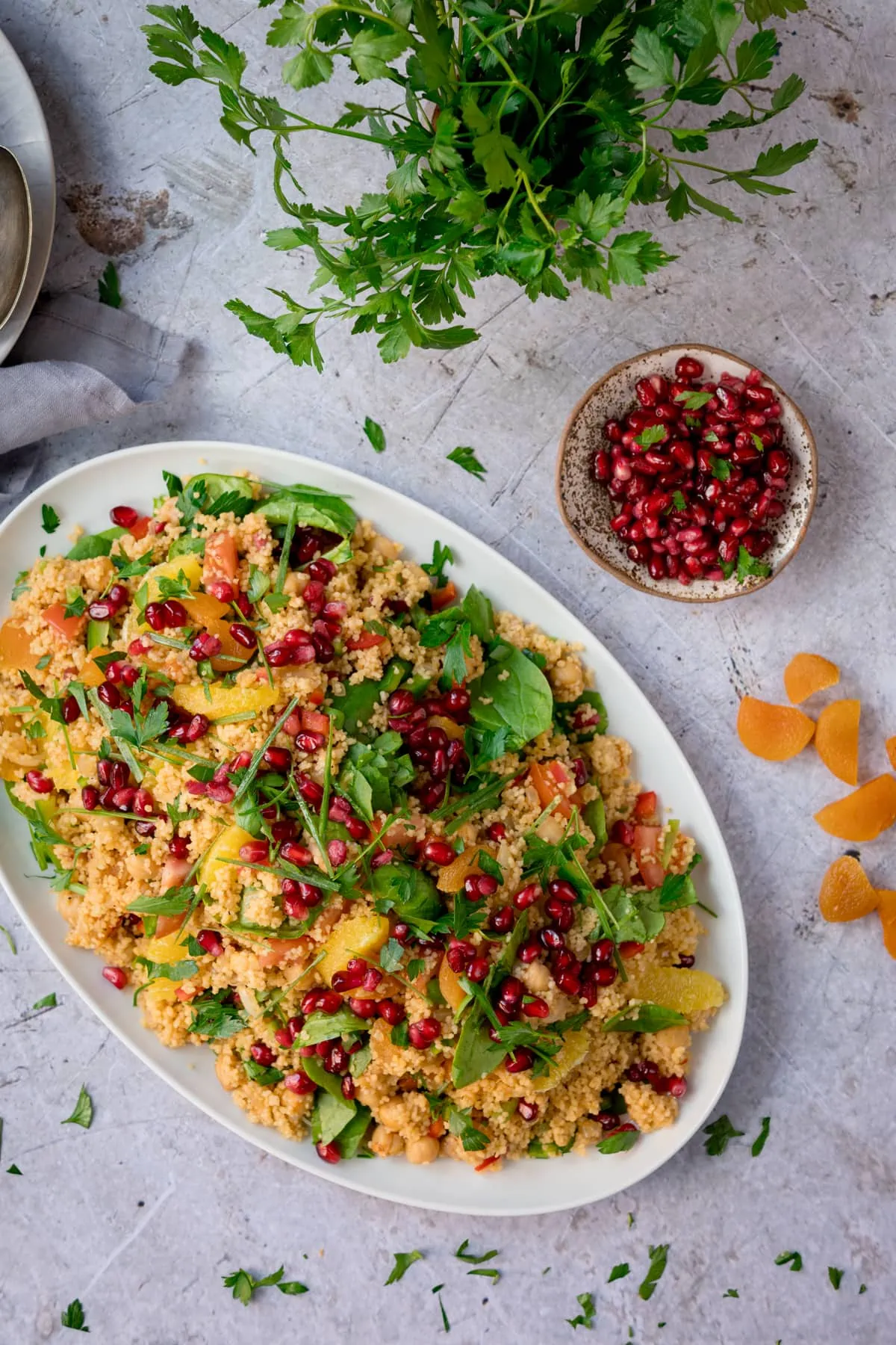 Overhead image of a large oval white plate filled with Moroccan style couscous with oranges and pomegranate. The plate is on a white surface next to a dish of pomegranate. There is chopped dried apricot and and fresh herbs scattered around.