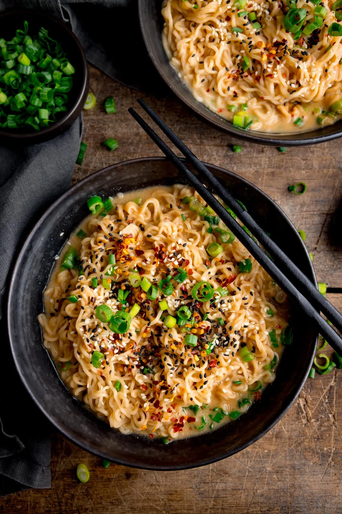 Overhead image of a black bowl filled with ramen noodles topped with chilli flakes, spring onions and mixed sesame seeds. There is a pair of black chopsticks resting on top of the bowl. The bowl is on a wooden table, next to a grey napkin, small bowl of chopped spring onions and a further bowl of noodles.