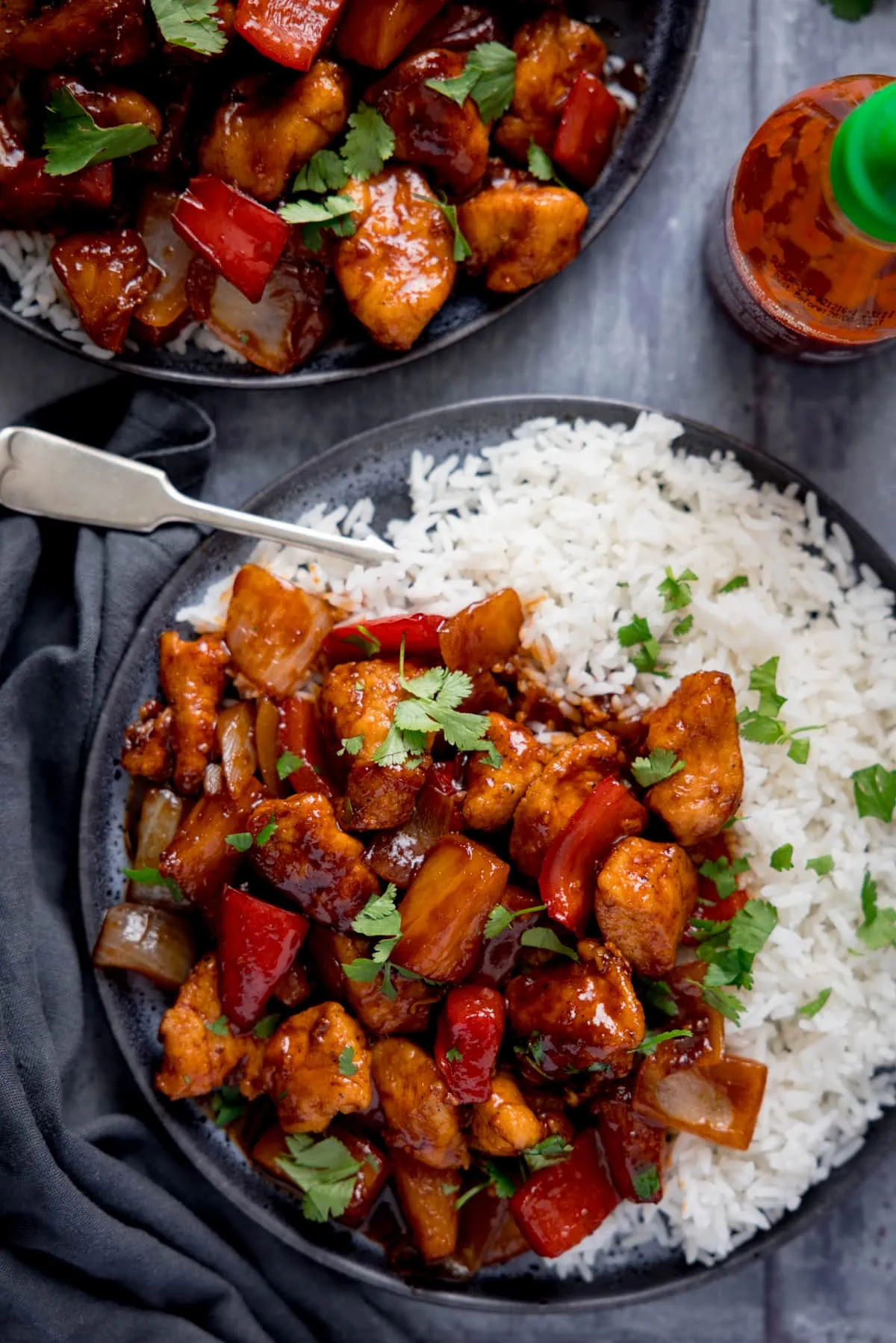 Overhead image of sticky pineapple chicken stir fry and boiled rice on a dark plate with a fork sticking out. The plate is on a blue-grey background, next to a blue-grey napkin. There is a further plate of stir fry and a bottle of sriracha sauce also in shot.