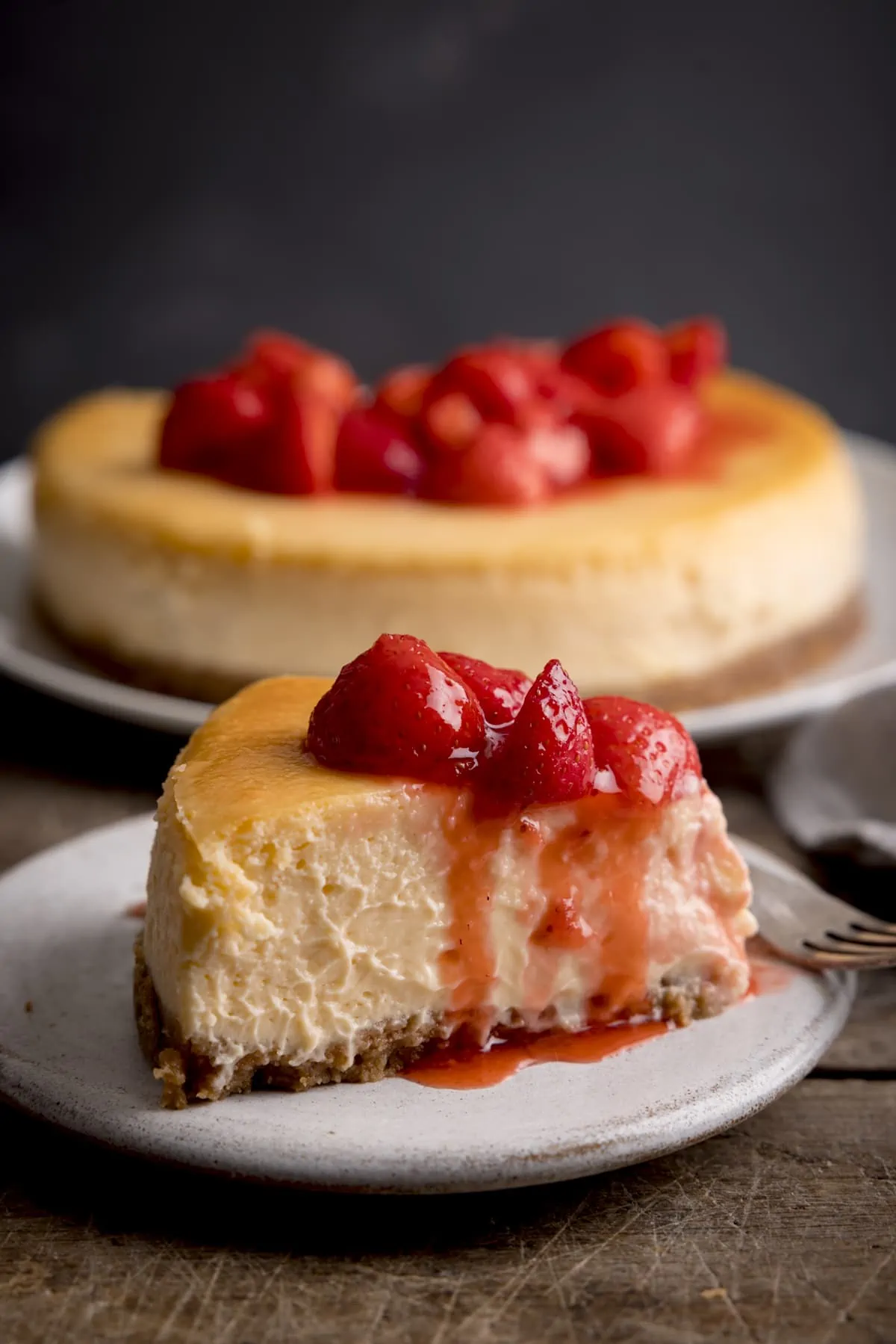 A close up of a slice of strawberry cheesecake on a white plate. The rest of the cheesecake is on a plate in the background.