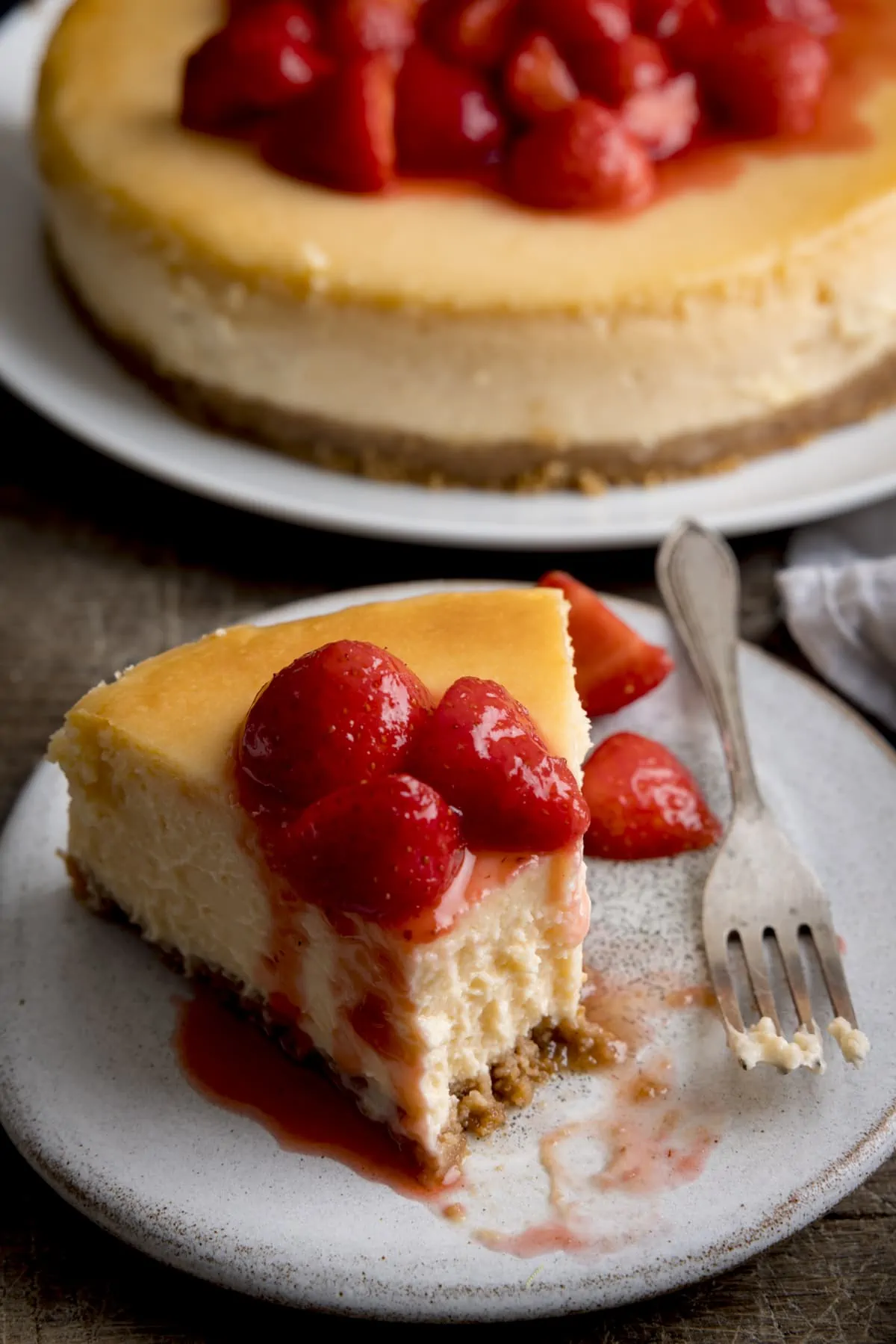 Close up of a piece of strawberry cheesecake on a white plate with a bite taken out. The rest of the cheesecake is on a plate in the background.