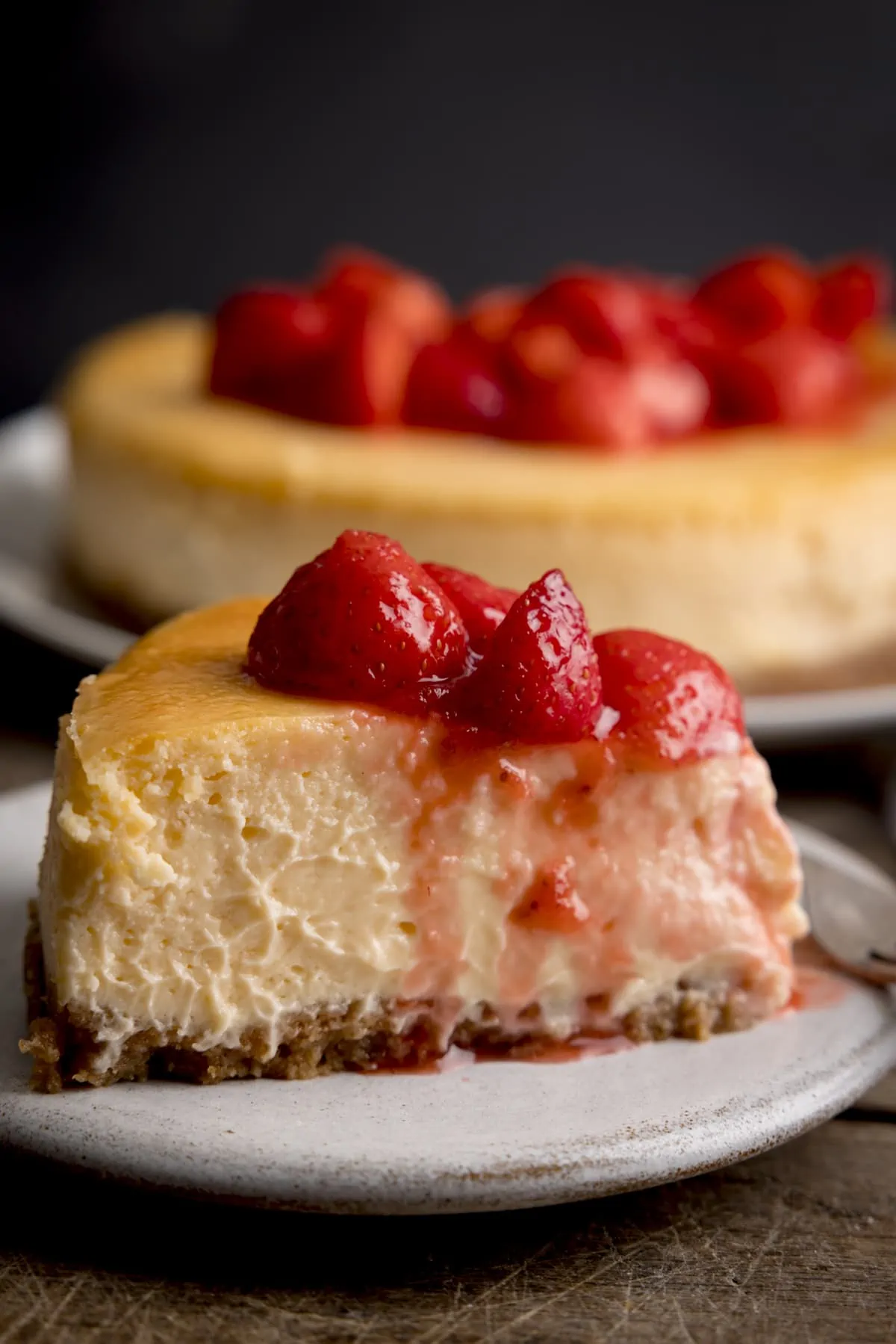 Side shot of a slice of strawberry cheesecake on a white plate against a dark background. The rest of the cheesecake is on a plate in the background.